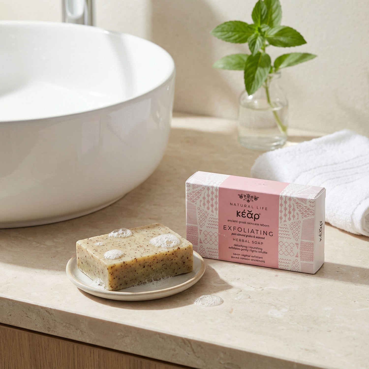 Bar of Kear Exfoliating Herbal Soap on a plate next to its packaging on a bathroom counter with a plant and towel in the background.