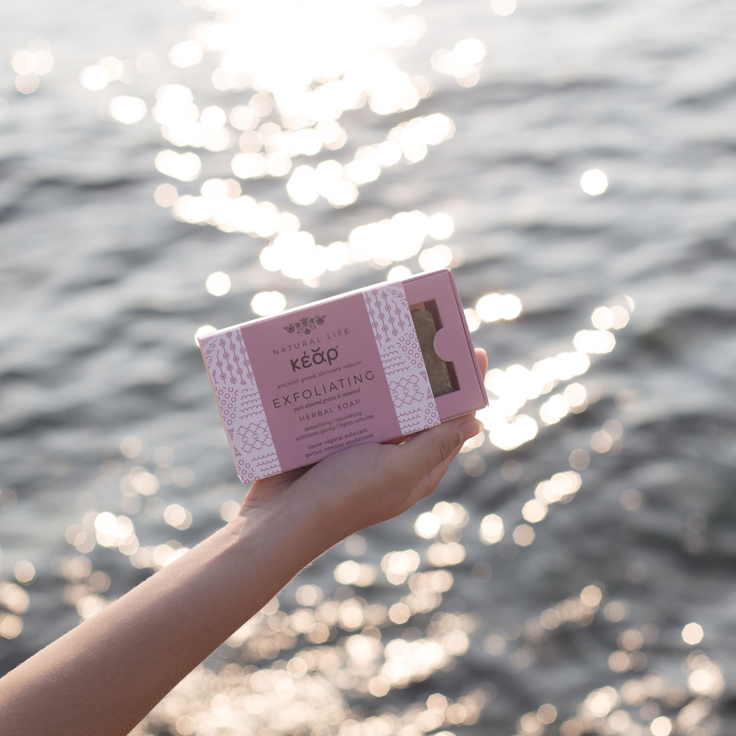 Hand holding Kear Exfoliating Herbal Soap with almond grains and seaweed extract against water background.
