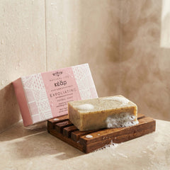 Bar of Kear Exfoliating Herbal Soap on a wooden soap dish with packaging in the background.