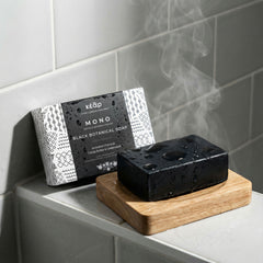 Kear MONO Black Botanical Soap with activated charcoal on a wooden soap dish on a bathroom tiled shelf.