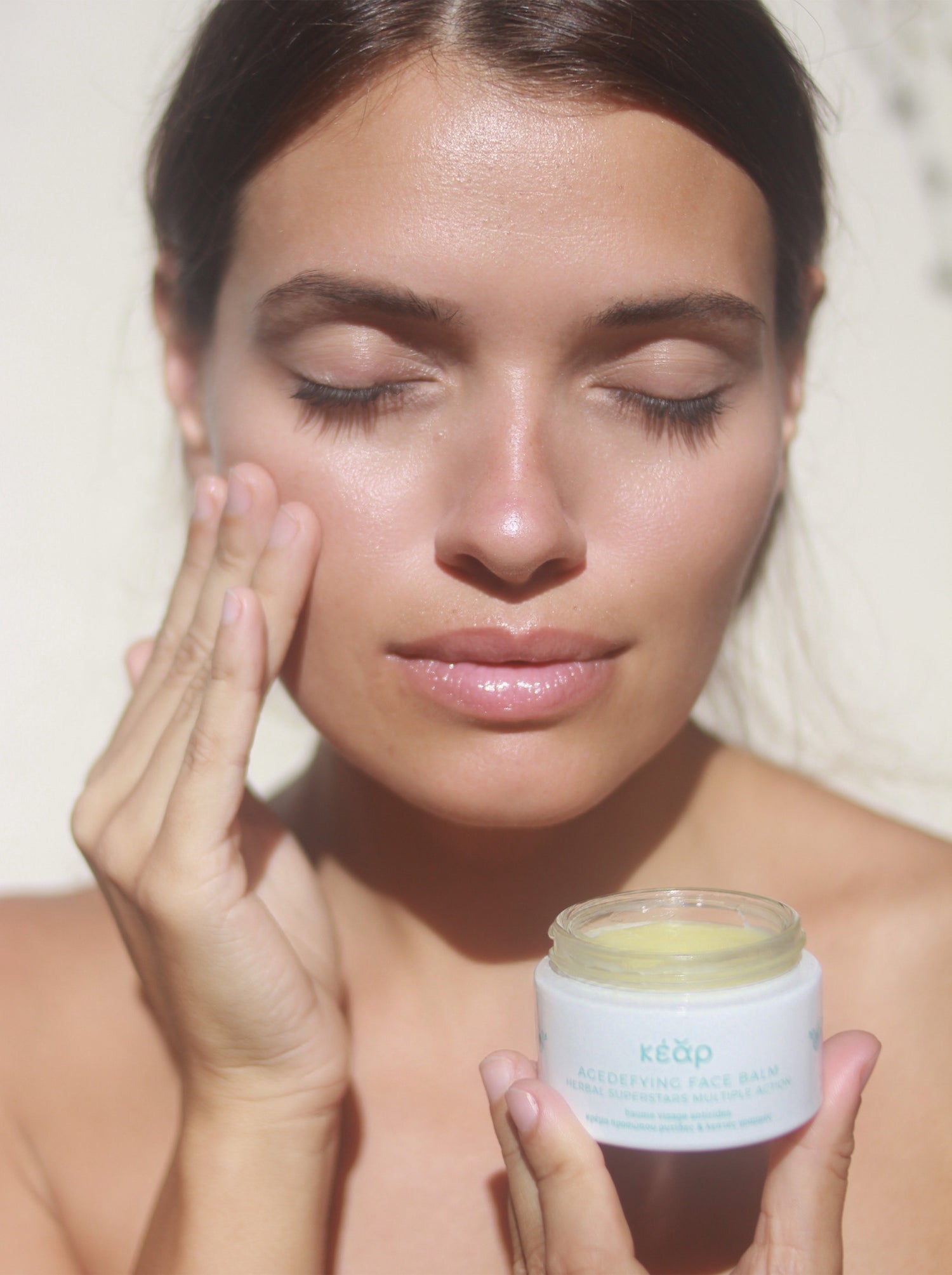 Woman applying Kear AgeDefying face balm on face and holding glass jar in warm sunlight.