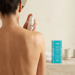 Person applying Kear Hydrate Relax natural body oil on shoulder, with a box of the same product in the background.
