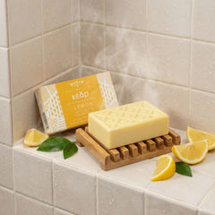 Kear natural Lemon soap bar on a wooden soap dish with lemon slices on a tiled bathroom shelf.