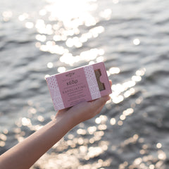 Hand holding Kear Exfoliating Herbal Soap with almond grains and seaweed extract against water background.