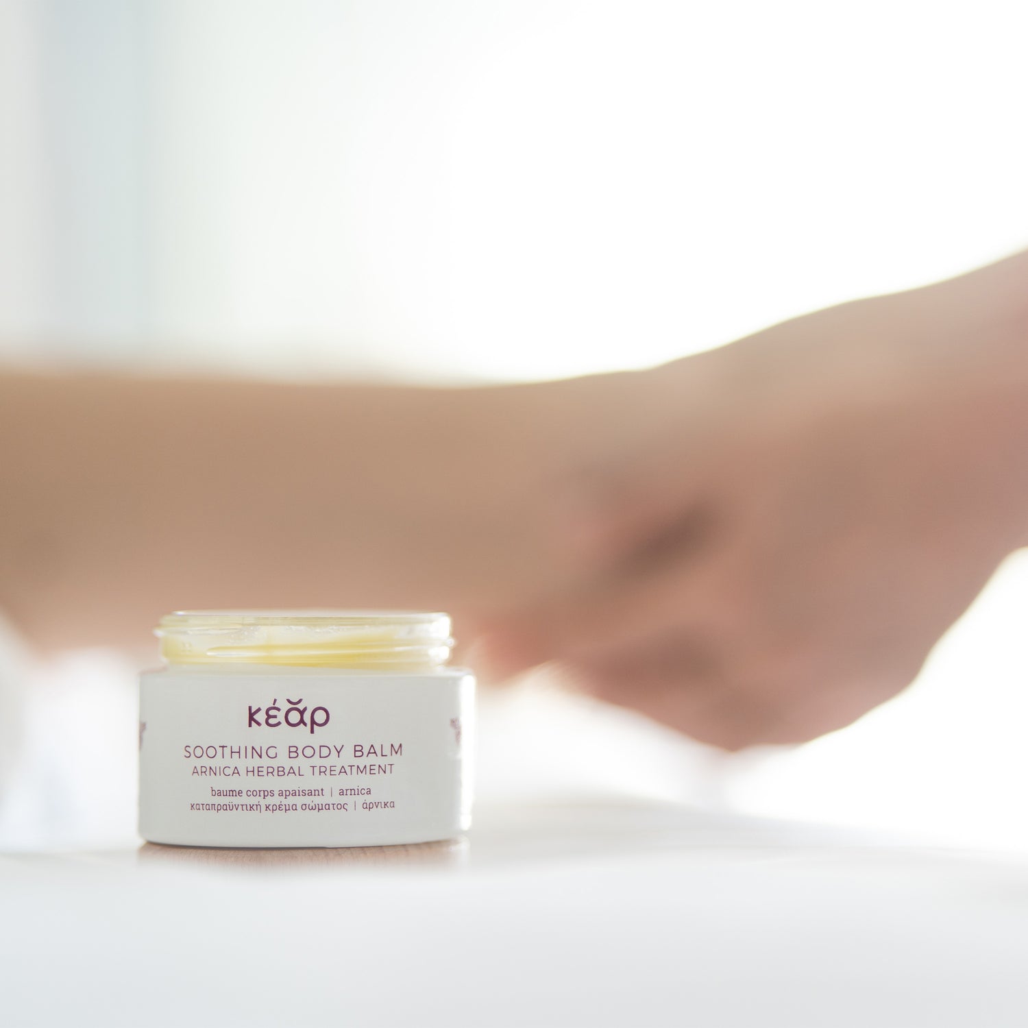 Open glass jar of Kear Soothing body balm with hands massaging leg in the background.