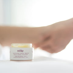 Open glass jar of Kear Soothing body balm with hands massaging leg in the background.