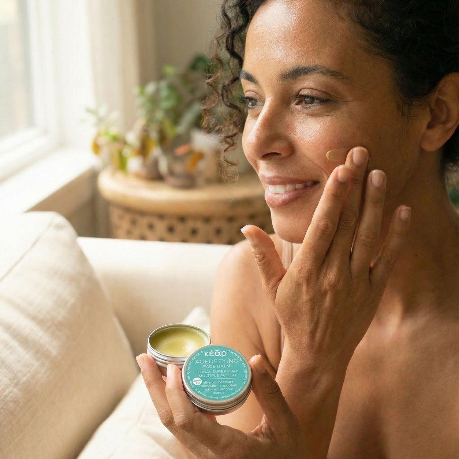 Woman applying Kear waterless AgeDefying face balm and holding the travel size jar in a cozy indoor setting.